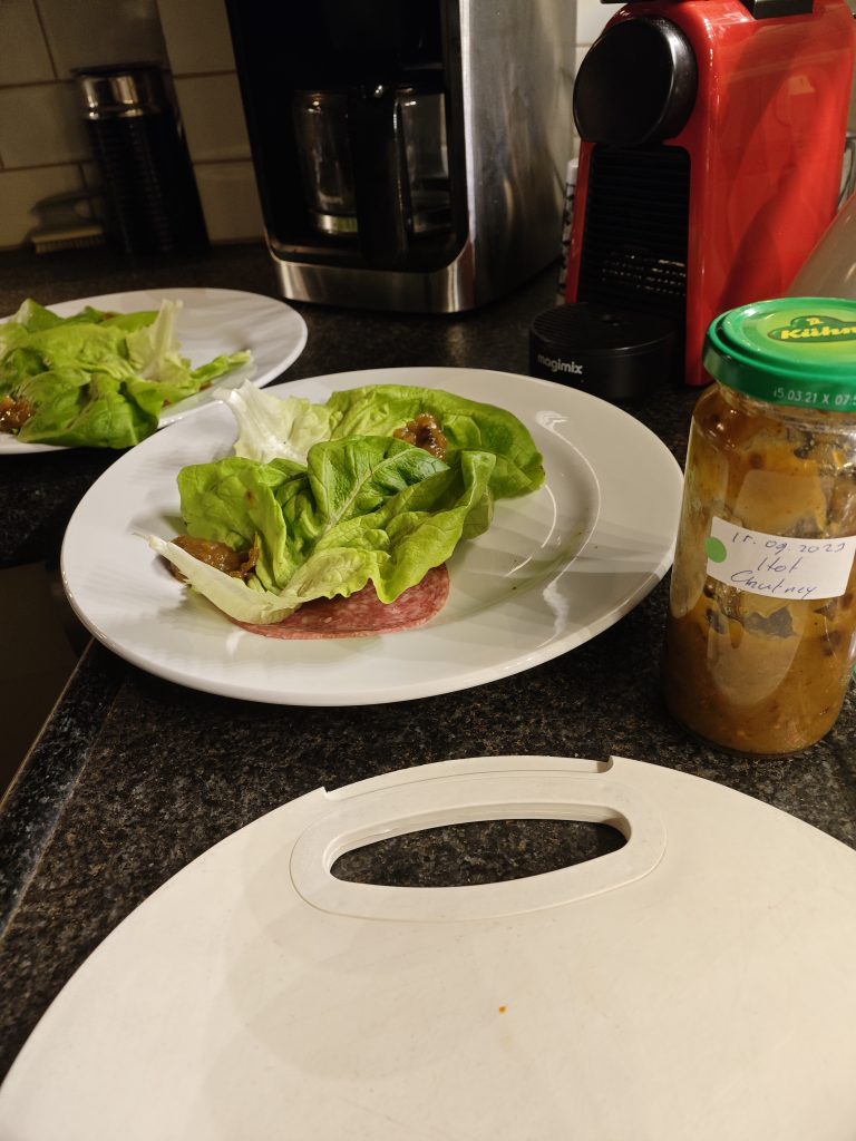 Two plates with green lettuce leaves and meat slices, a jar of chutney labeled with a date, and a red coffee machine on a kitchen counter.
