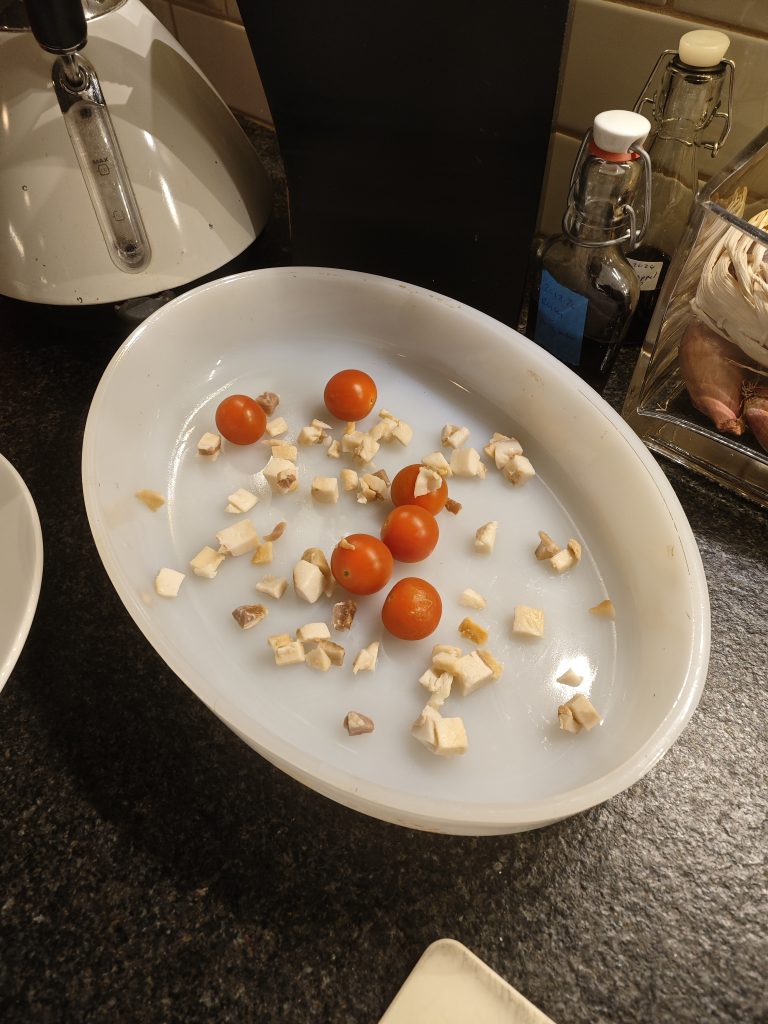 Alt text: Oval dish containing cherry tomatoes and diced mushrooms on a dark countertop. Nearby are bottles with swing-top lids and a white appliance.