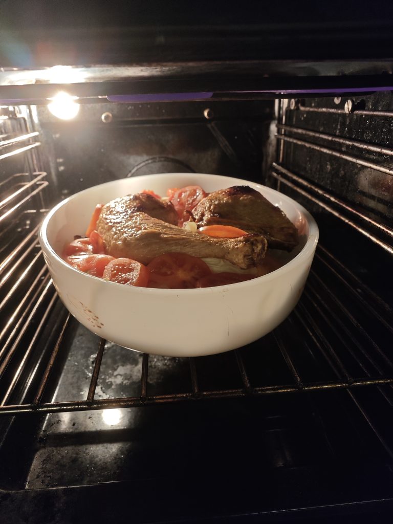 Roasting dish inside an oven.

Two chicken leg quarters on top.

Slices of red tomatoes visible.

Round white ceramic baking dish.

Oven interior with metal racks and a light source.