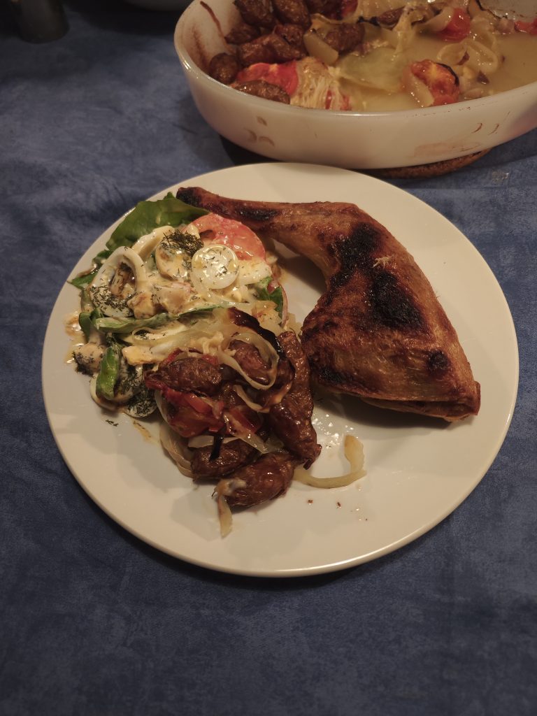 Plate with cooked chicken leg, browned skin.

Salad with sliced tomatoes, cucumber, and lettuce.

Dill sprinkled on salad; white dressing visible.

Roasted potatoes and onions on plate.

Casserole dish in background with browned meat and vegetables, including tomatoes and onions.

Blue tablecloth.