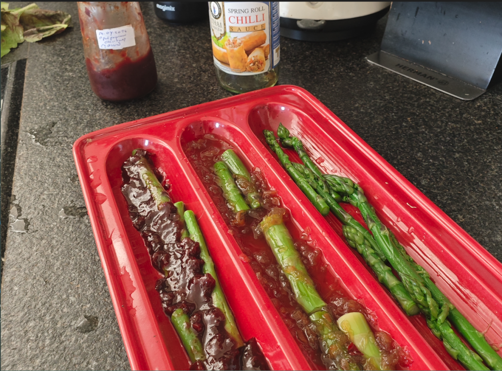 Red tray featuring asparagus spears spread across compartments.; Dark leafy greens placed under a layer of sauce in the left compartment.; Glass jar on the left with a handwritten label dated 15-07-2023.; Bottle of "Thai Dragon Spring Roll Chilli Sauce" in the background.; Black countertop surface, partially visible kitchen appliances.