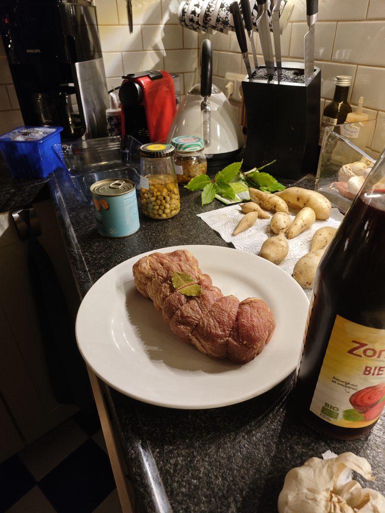 A kitchen counter with a tied roast on a white plate next to potatoes, a can of peaches, a jar of olives, and a bottle of beer, surrounded by various kitchen appliances and utensils.