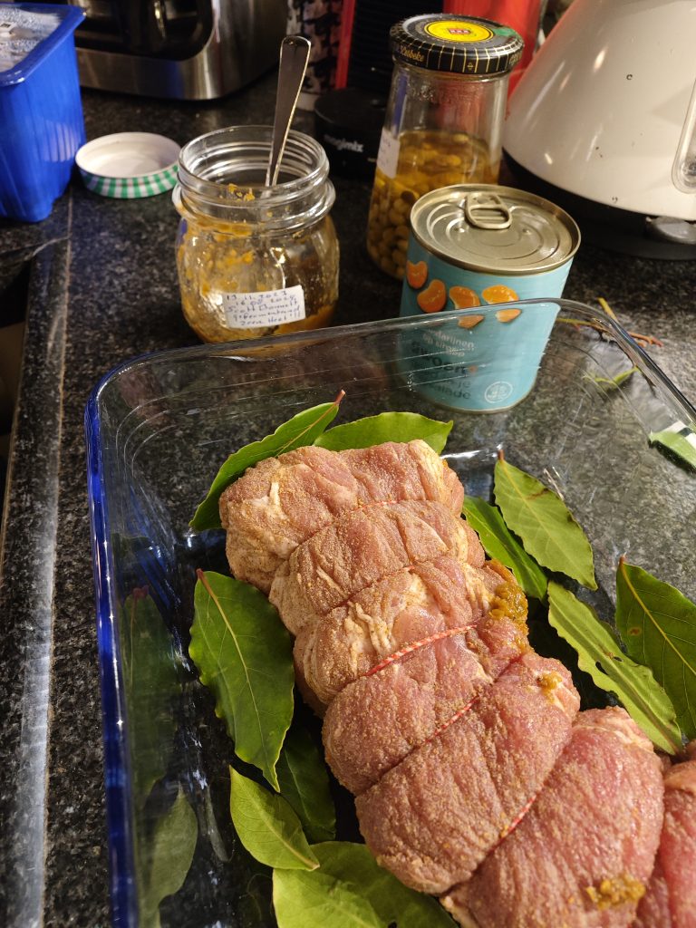 A tied raw pork tenderloin seasoned and placed on bay leaves in a glass baking dish, with an open jar of mustard and a tin of mandarin oranges nearby on a kitchen countertop.