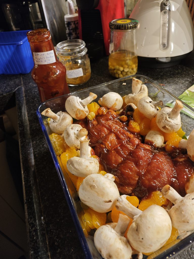 A baking dish on a kitchen counter filled with a glazed roast surrounded by whole mushrooms and pieces of fruit, next to a bottle of sauce and various jars.