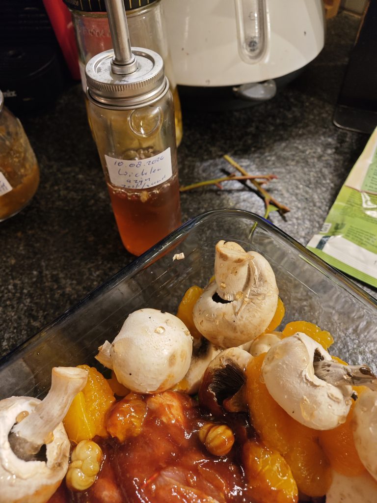 A glass dish containing whole mushrooms, yellow fruit slices, and a red sauce on a kitchen counter. In the background, a bottle with a pump dispenser and handwritten label, along with other kitchen items, are visible.