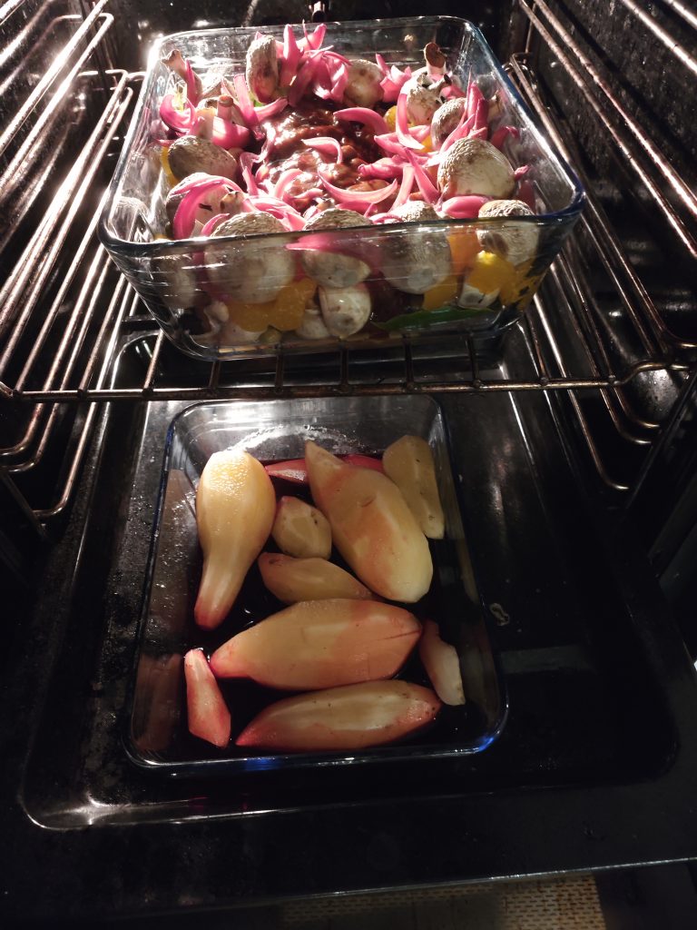 Two glass baking dishes in an oven: the top dish contains mushrooms, sliced red onions, tomatoes, and bell peppers; the bottom dish contains peeled red-skinned fruits.