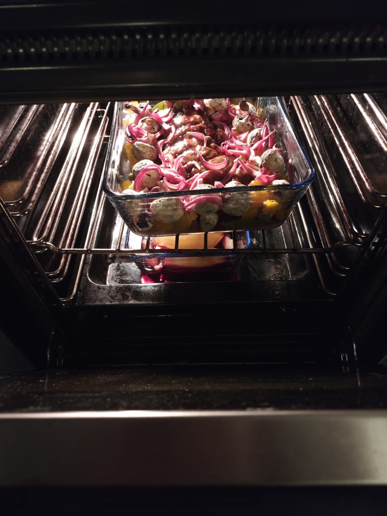 A glass baking dish in an oven containing a mix of vegetables, mushrooms, and red onions.