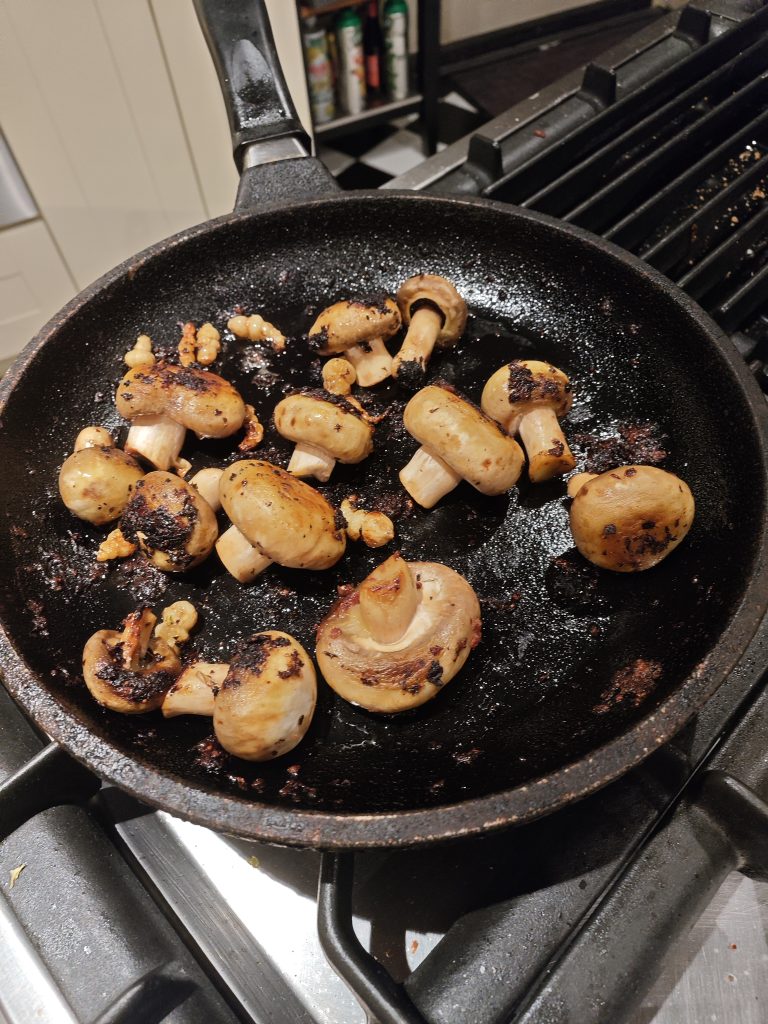 A frying pan on a stove containing sautéed mushrooms and small pieces of charred garlic or seasoning.